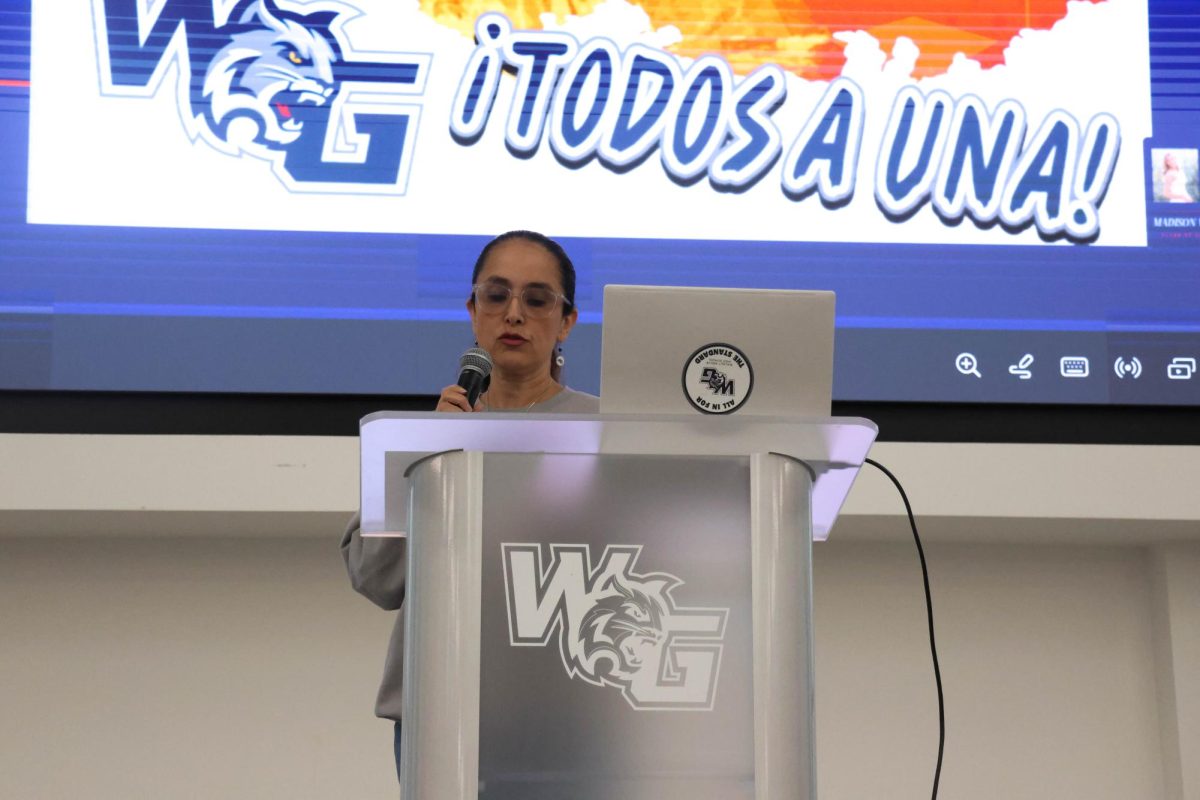 Mrs. Mora gives a speech at the start of the ceremony to honor the Seniors in Spanish Honors Society. The slide in the back has the society's motto "Todos a una" (all as one).
